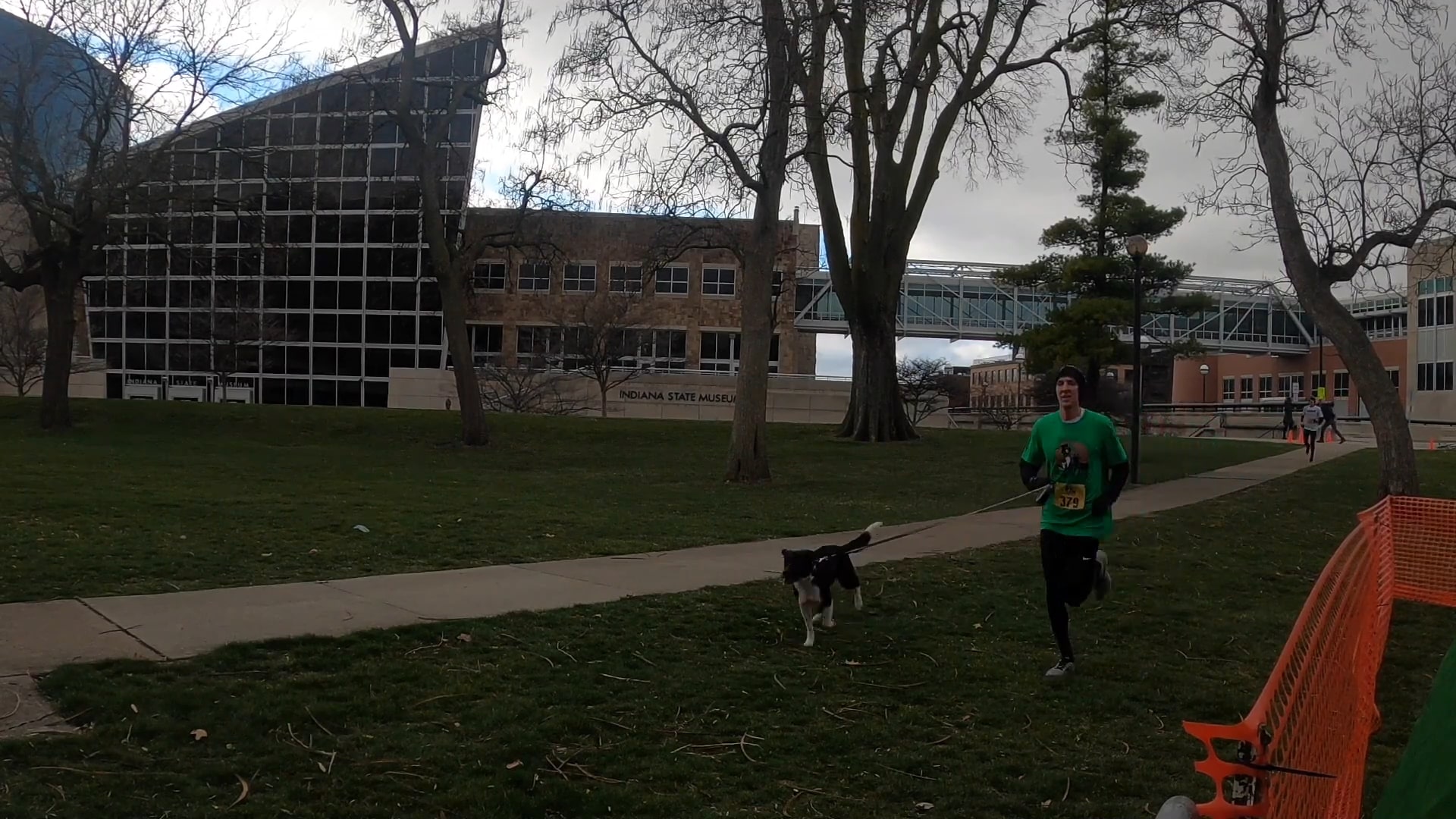 A runner wearing a green shirt and race bib jogs alongside their black and white dog on a pathway near the Indiana State Museum during a 5K race