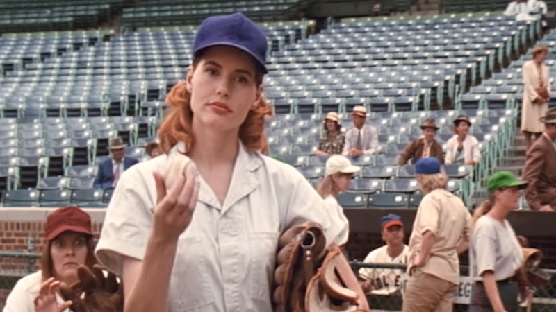 A female baseball player stands on the field in uniform, surrounded by teammates and extras at the stadium in A League of Their Own
