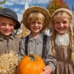 Three smiling children in rustic clothing holding a pumpkin at a festive autumn celebration with pumpkins and hay bales
