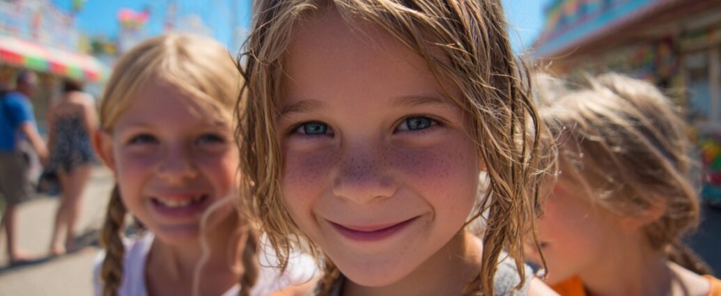 Three smiling children at an outdoor fair with colorful booths and rides in the background
