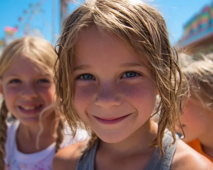 Three smiling children at an outdoor fair with colorful booths and rides in the background