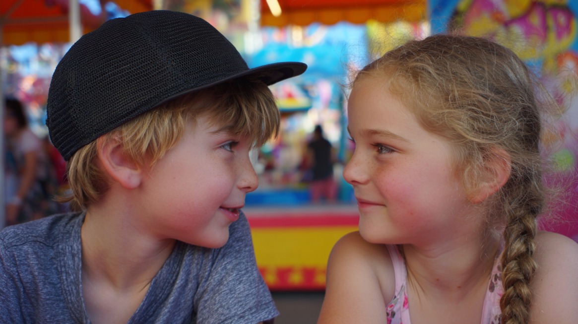 A young boy in a black cap and a girl with braided hair smiling at each other at a colorful fairground