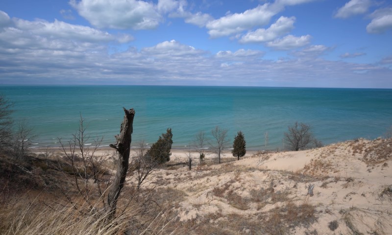 A tranquil beach scene with grassy dunes leading to a turquoise lake under a partly cloudy sky