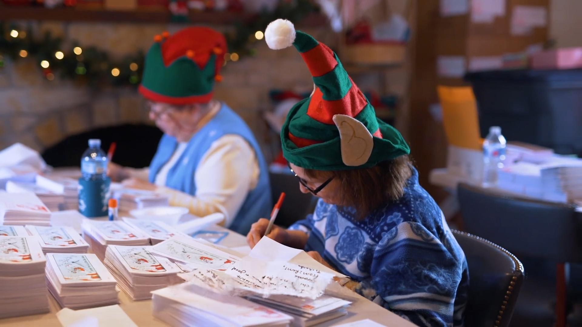 Volunteers wearing elf hats handwrite replies to children's Santa letters at a festive workspace