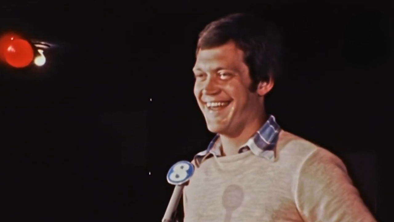 David Letterman smiles beside a microphone on a dark stage