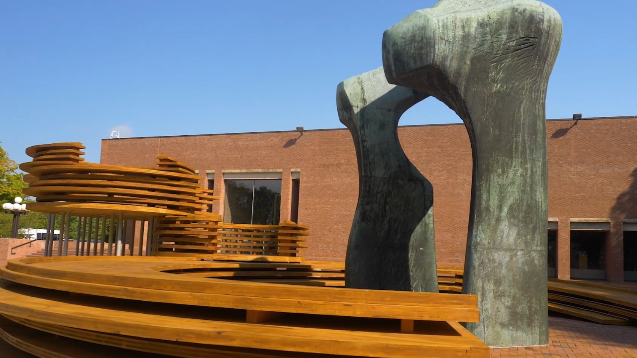 A large bronze arch sculpture stands beside curved wooden benches in a brick plaza