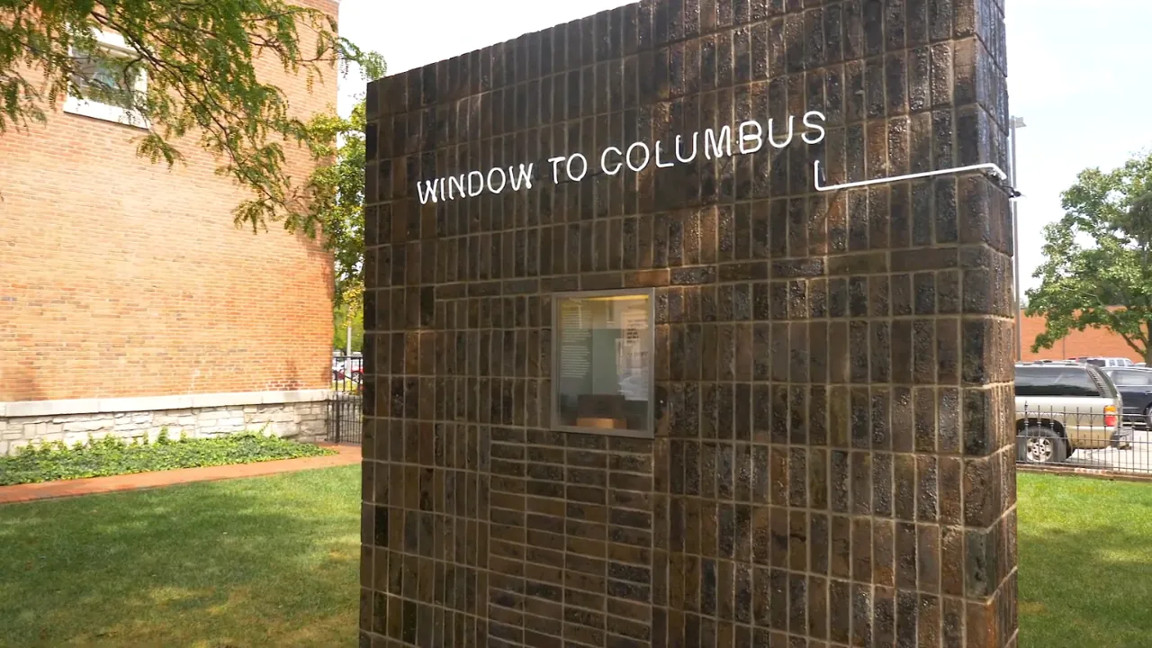 A dark tile art wall labeled Window to Columbus stands on a grassy downtown site